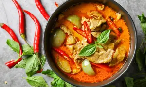 Miseczka czerwonego curry z mięsem, warzywami i papryczkami chili, otoczona świeżymi ziołami i papryczkami.
