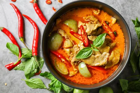 Miseczka czerwonego curry z mięsem, warzywami i papryczkami chili, otoczona świeżymi ziołami i papryczkami.