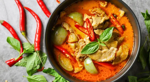 Miseczka czerwonego curry z mięsem, warzywami i papryczkami chili, otoczona świeżymi ziołami i papryczkami.