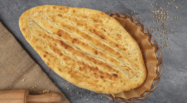 Chlebek naan w koszyku, obok wałek i ziarna pszenicy.